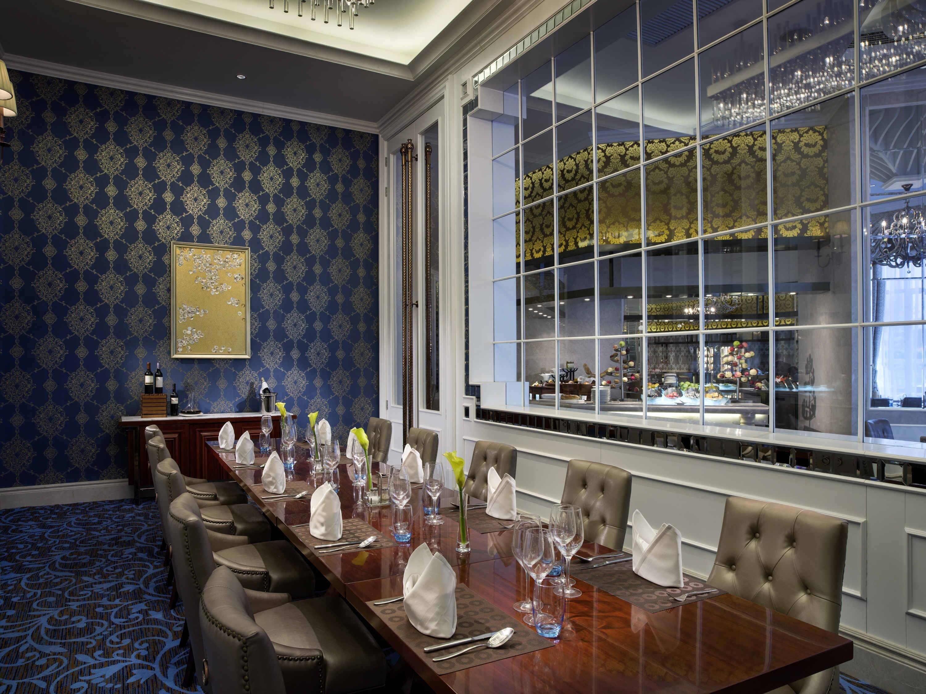 A conference room with a long table set for a meeting, a large window with a view of a restaurant, and a patterned wallpaper.