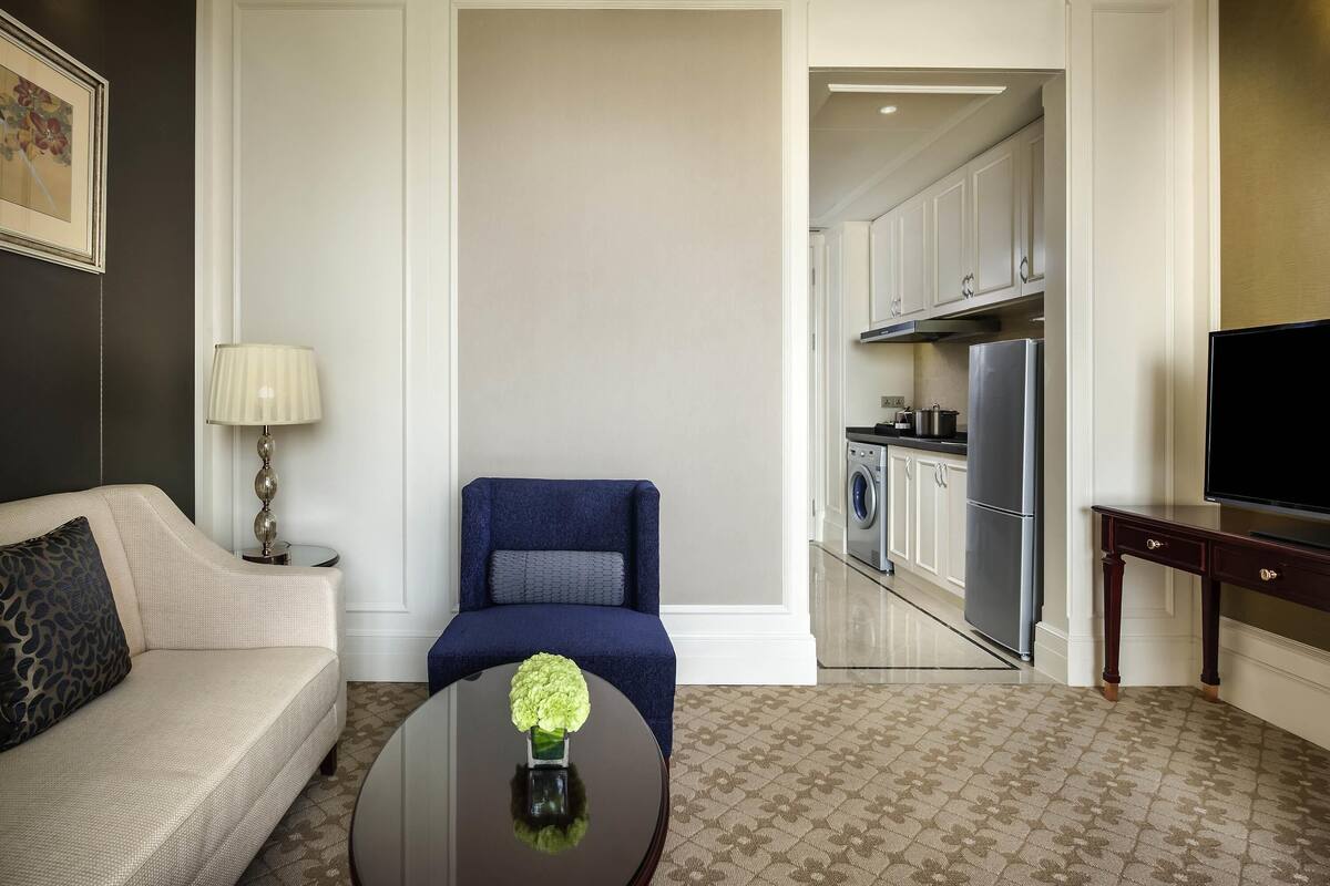 A modern hotel room with a sofa, a blue armchair, a coffee table with a vase, a TV, and a kitchen area visible through an open door.