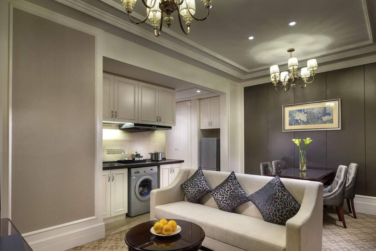 A living room with a sofa, a coffee table, a dining area with chairs, a chandelier, and a kitchen with appliances.