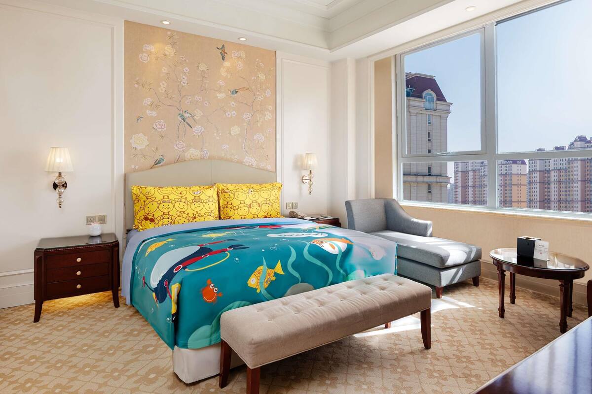 A hotel room with a large bed, a bench, a chair, a small table, and a view of the city through a large window.