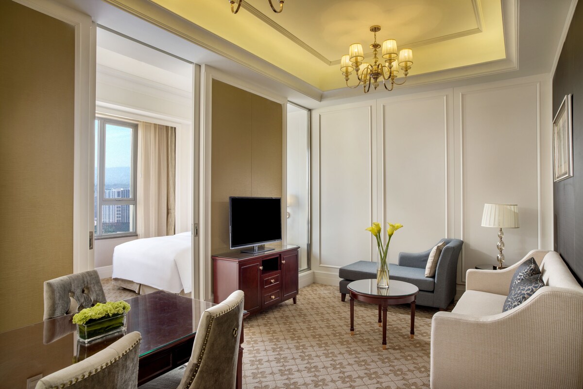 Habitación de hotel con cama, televisor, escritorio, sillón, sofá y una pequeña mesa con flores.