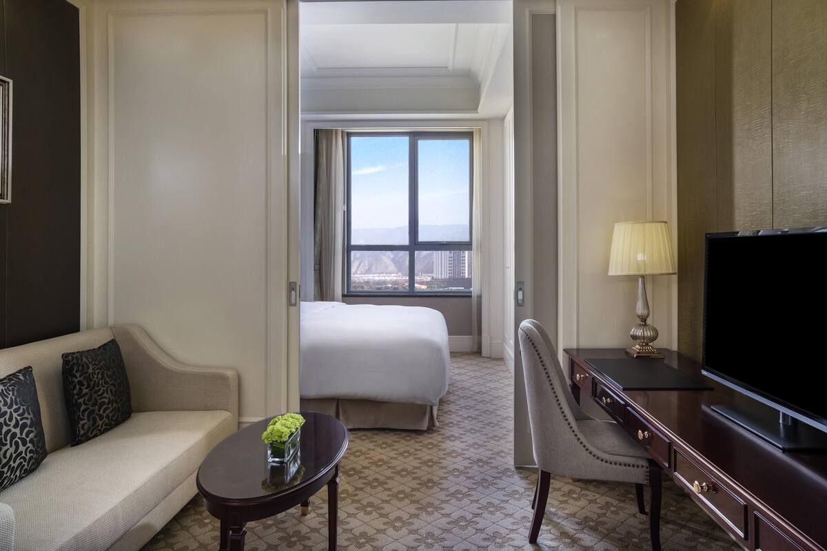 A hotel room with a bed, a sofa, a desk, a television, and a window with a city view.