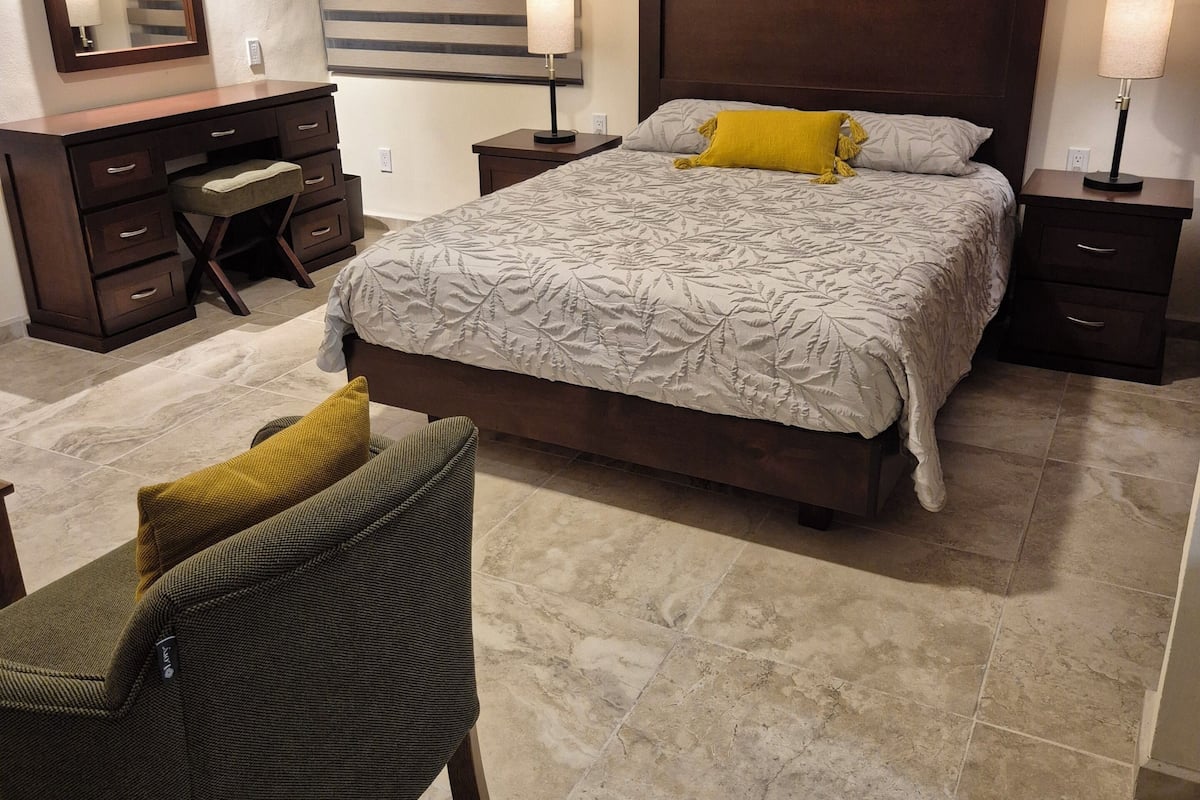 Un dormitorio con cama, mesitas de noche, cómoda, silla, espejo y ventana con persianas.