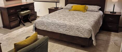 Un dormitorio con cama, mesitas de noche, cómoda, silla, espejo y ventana con persianas.