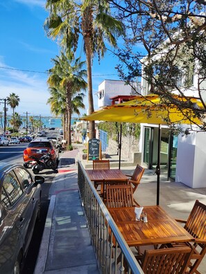 Ein Café am Meer mit Außenbereich, Schild mit Kaffee und Frühstücksangeboten, sowie geparkte Autos entlang der Straße.