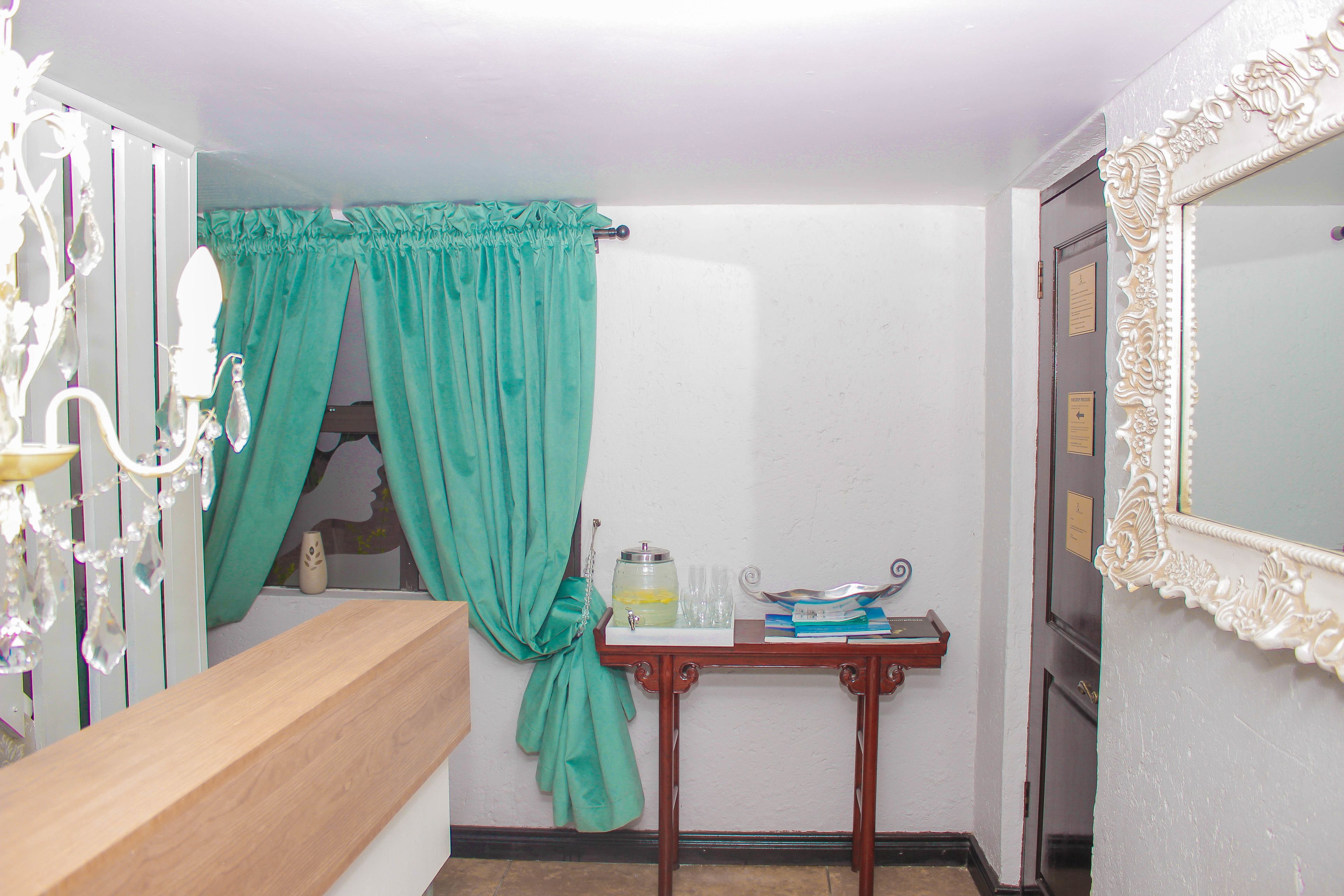Une salle de bain avec un comptoir en bois, un rideau vert, un miroir doté d’un cadre orné et une petite table sur laquelle se trouvent une carafe et des verres.