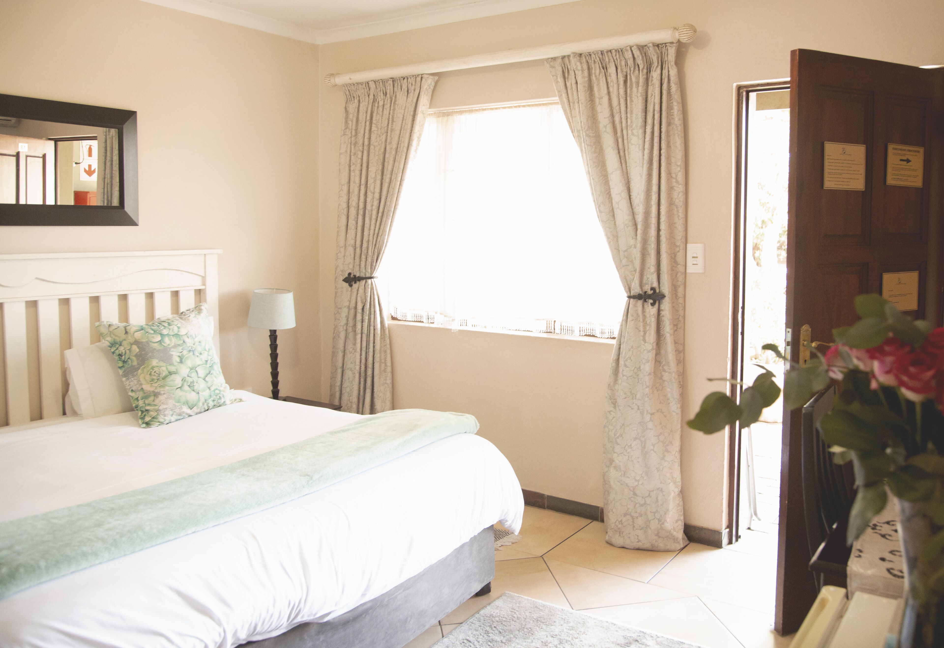A bedroom with a bed, a bedside lamp, a window with curtains, and a door with a sign.