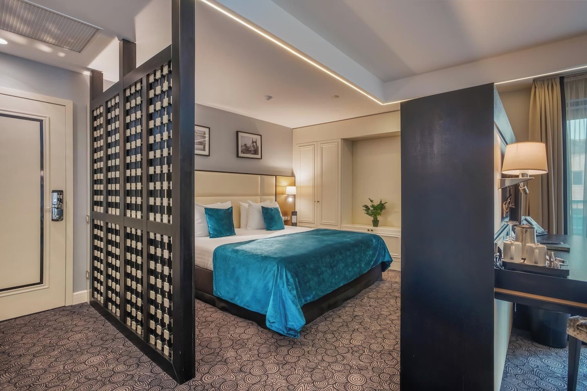 A hotel room with a bed, a desk, a chair, a lamp, a plant, and a wall with a patterned design.