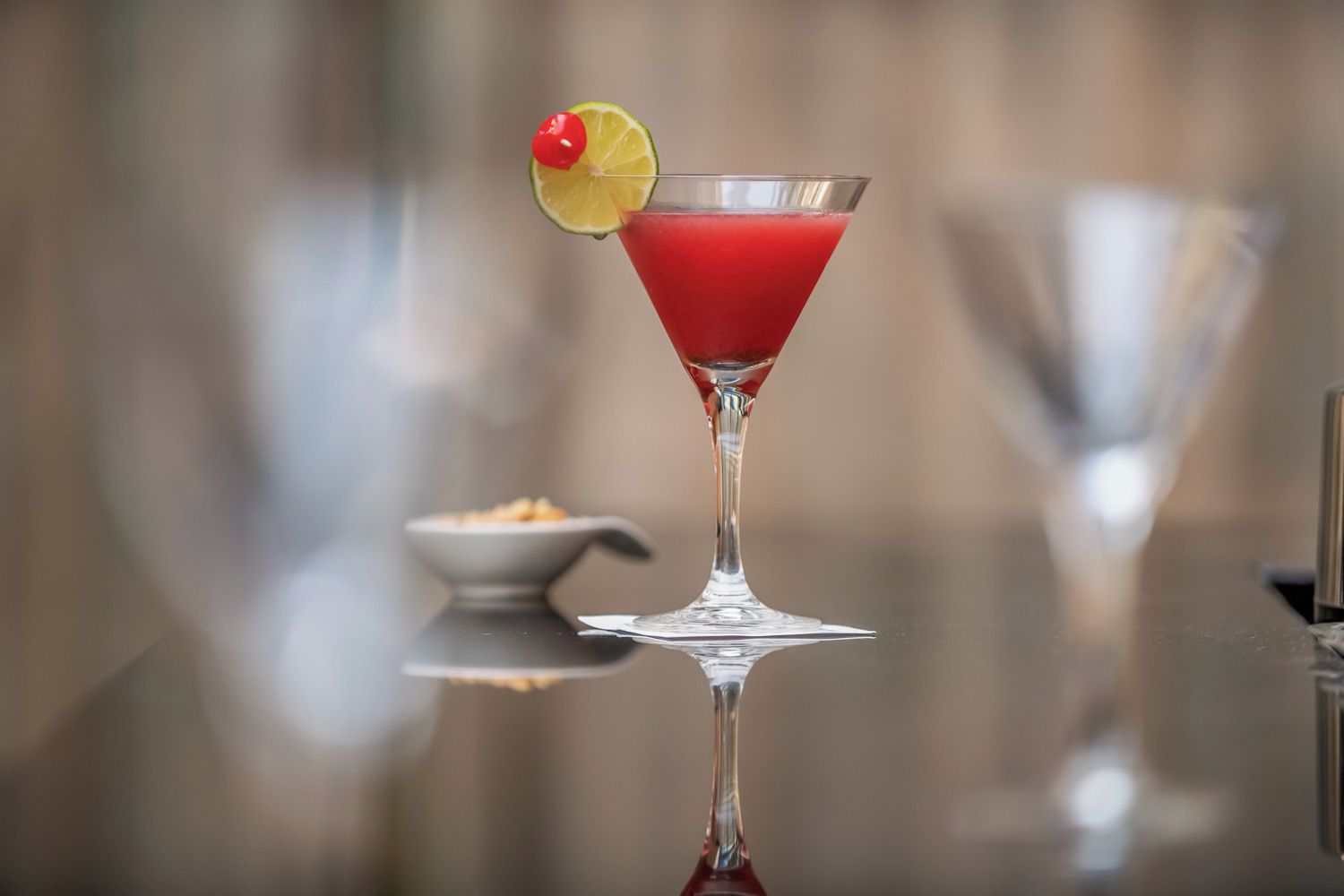 A martini glass with a red cocktail, garnished with a cherry and a slice of lime, on a reflective surface.