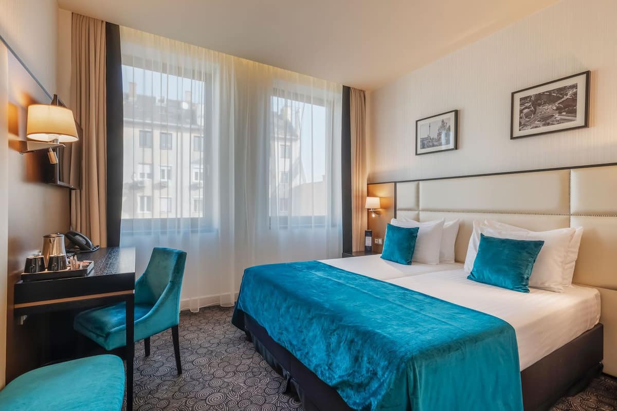 A hotel room with a bed, a desk, and a chair. There are two windows with sheer curtains, a wall-mounted lamp, and a framed picture on the wall.