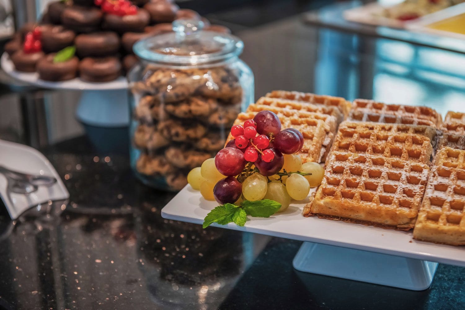 A buffet table with waffles, a jar of cookies, and a plate of grapes.