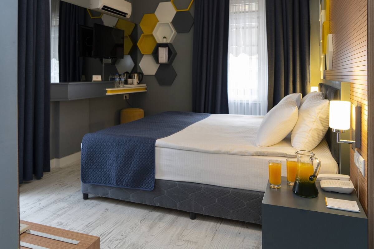 A modern hotel room with a large bed, a bedside table with a lamp, a small desk with a phone and a notepad, a mirror, a TV, and a window with curtains.