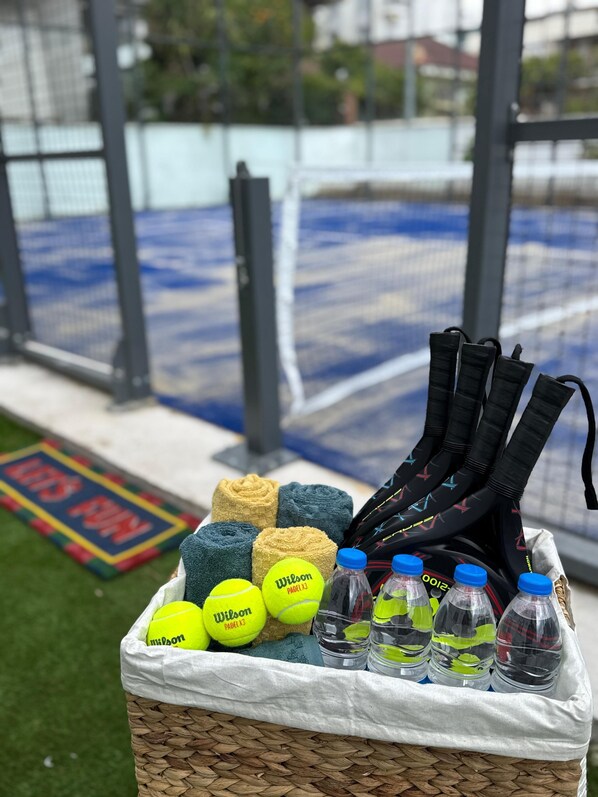 Una cesta con pelotas de tenis, una toalla y botellas de agua sobre un césped con una cancha de tenis al fondo.