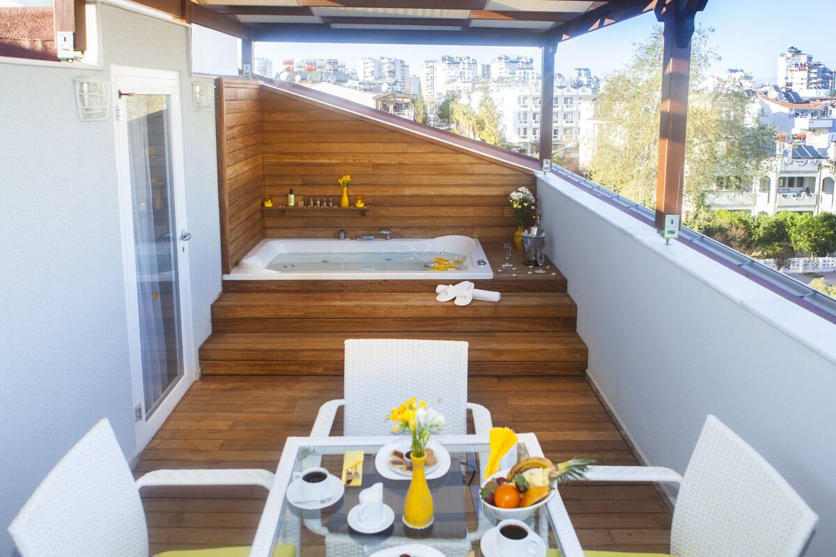 A balcony with a wooden floor, a glass table with breakfast items, a white chair, and a view of the city.