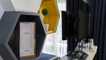 Habitación de hotel con un diseño moderno, que incluye un mueble hexagonal con un menú y una planta pequeña. Hay un televisor de pantalla plana montado en la pared, un escritorio con un teléfono y un control remoto, y una ventana con cortinas.