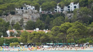 A coastal area with beach facilities, including umbrellas and lounge chairs, and buildings with satellite dishes on the hillside. - Alta Galdana (Ciutadella de Menorca)