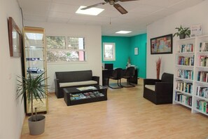 Ein Zimmer mit einer Couch, Sesseln, einem Couchtisch mit Zeitschriften, einem Bücherregal, einem Schreibtisch und einem Computer.