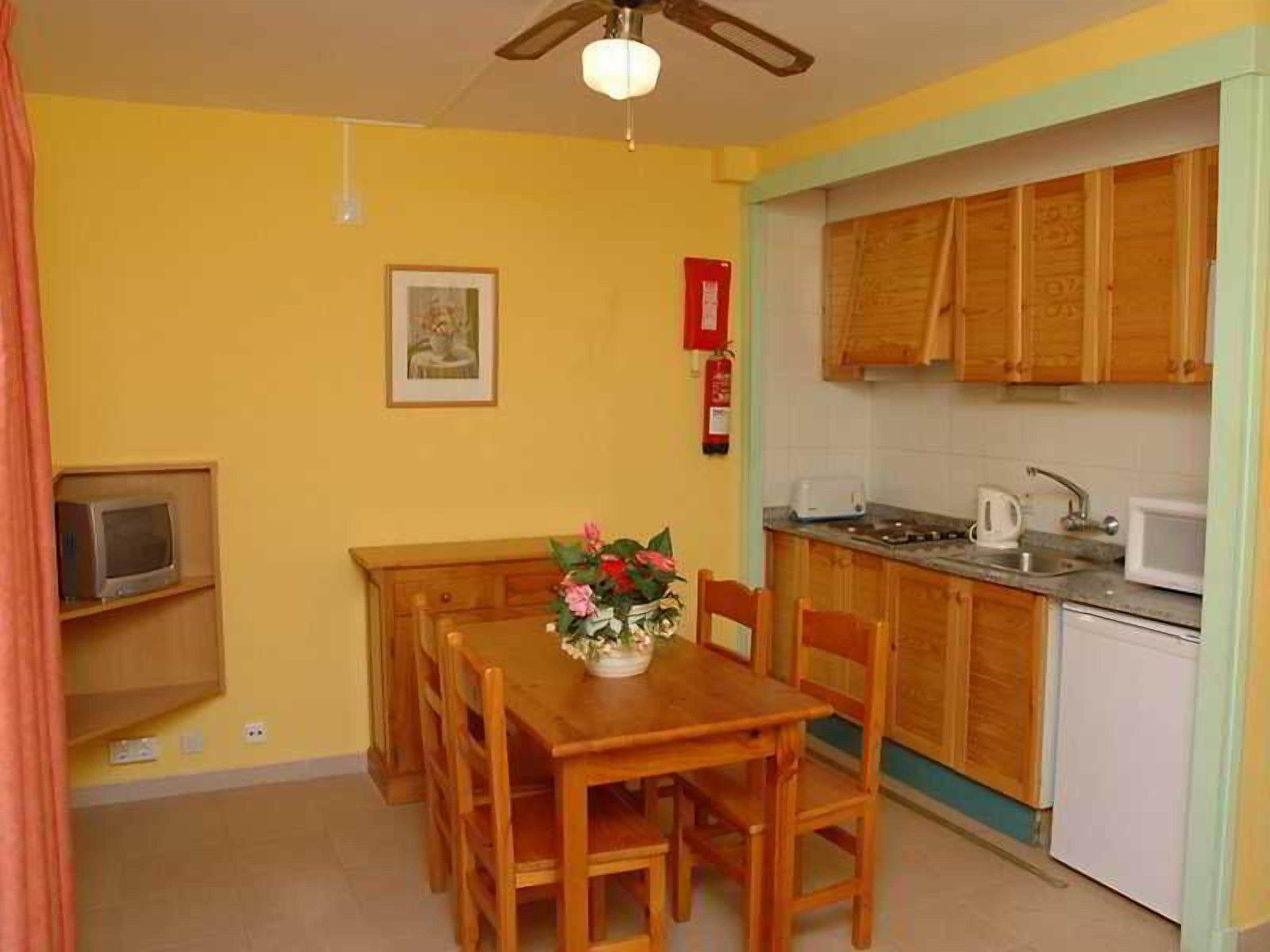 Une cuisine compacte avec des meubles en bois, une table à manger avec des chaises, une petite télévision et un ventilateur de plafond.