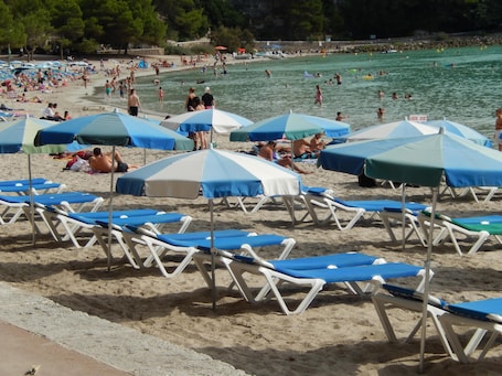 A beach with rows of sun loungers and umbrellas, a pool area, and people swimming and relaxing.