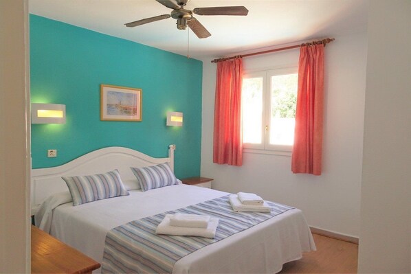 Ein Schlafzimmer mit Bett, Deckenventilator, Fenster mit Vorhängen und Wandleuchten.