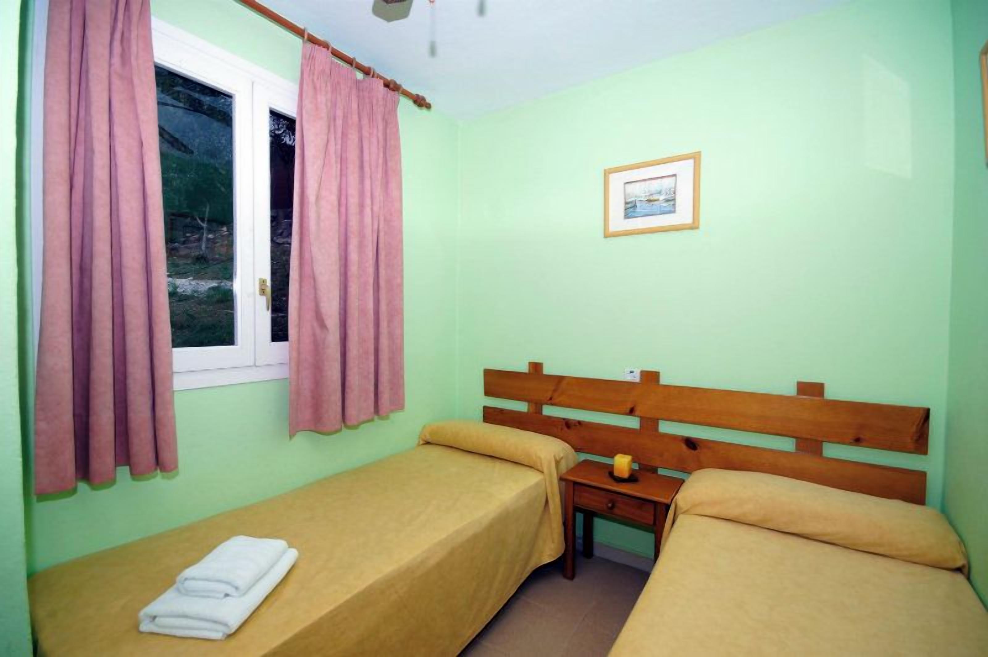 Una habitación con dos camas individuales, cabecero de madera, una mesita pequeña y una ventana con cortinas rosadas.