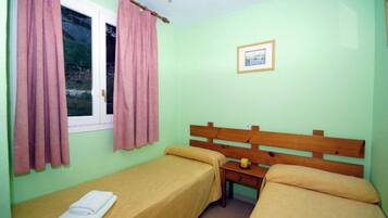 Una habitación con dos camas individuales, cabecero de madera, una mesita pequeña y una ventana con cortinas rosadas.