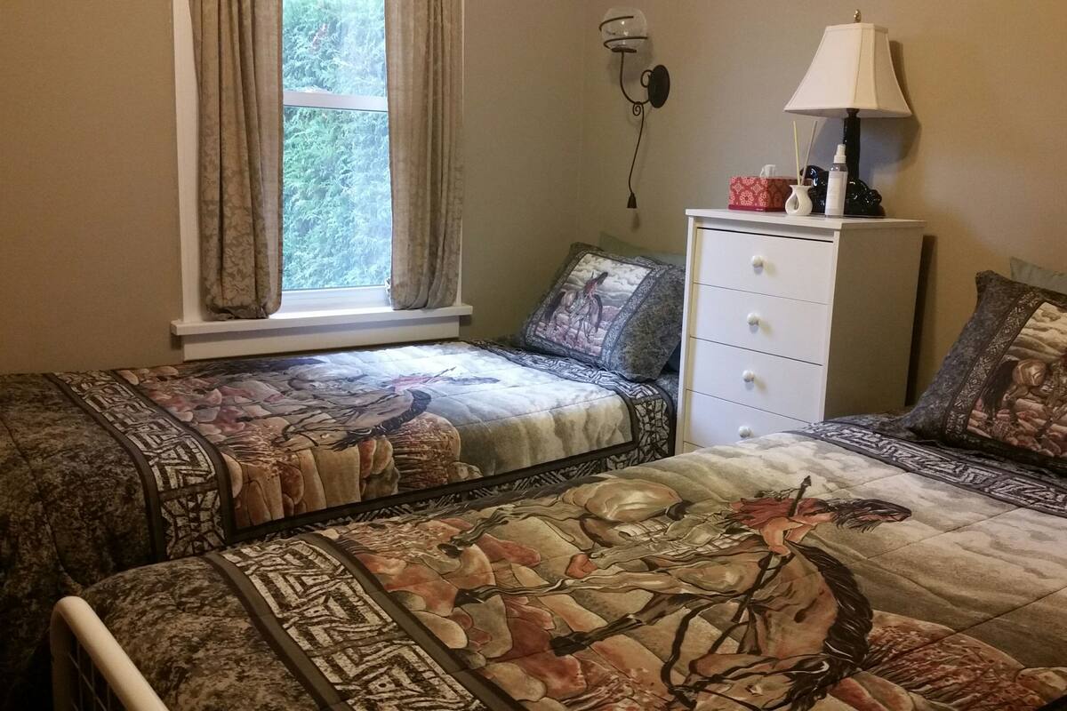 A bedroom with two beds, a window with curtains, a wall-mounted lamp, a bedside table with a lamp, and a dresser with drawers.