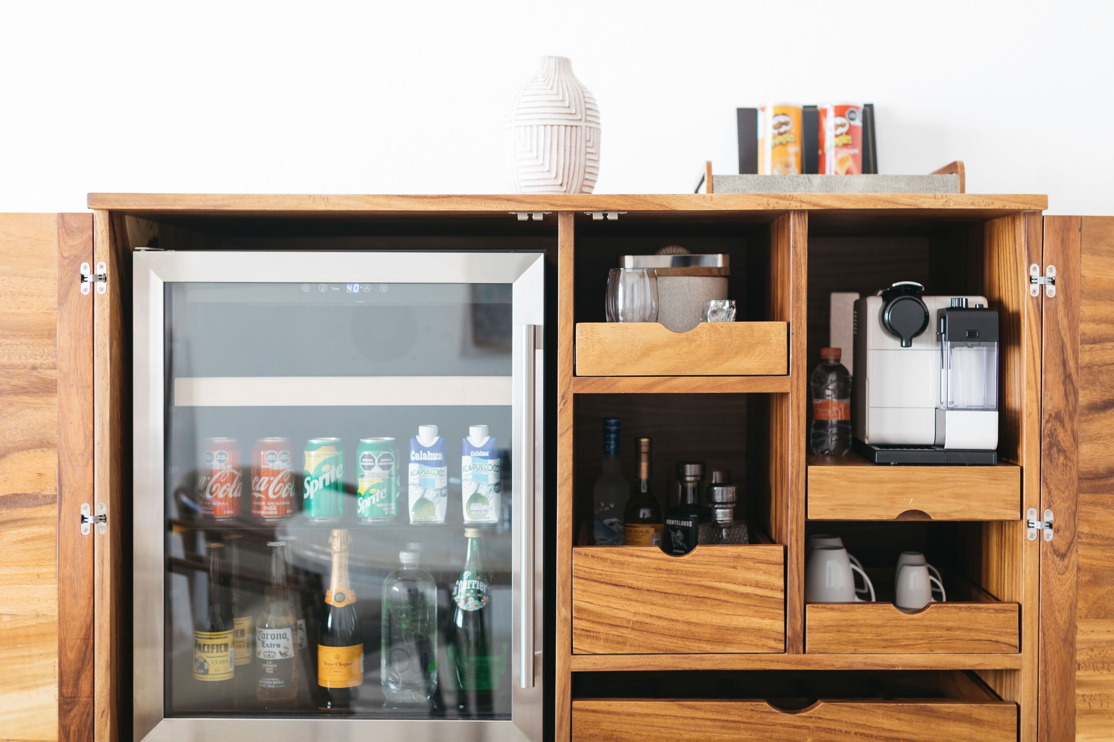 Un mueble de madera con puerta de vidrio que contiene bebidas y una máquina de café.