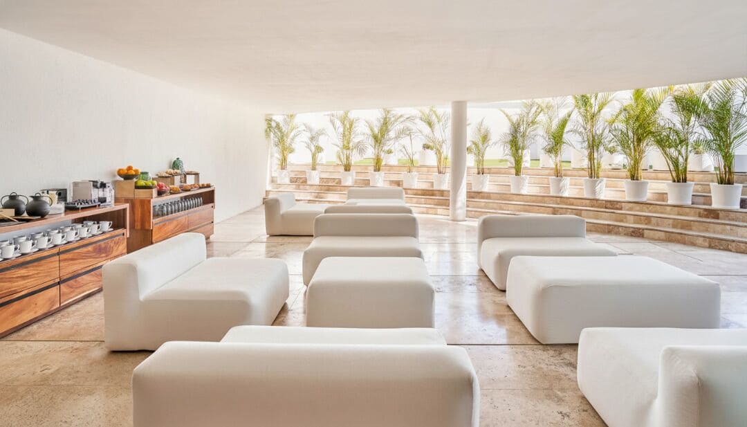 A modern lounge area with white sofas arranged in rows, a wooden sideboard with kitchenware, and a view of a landscaped garden with palm trees.
