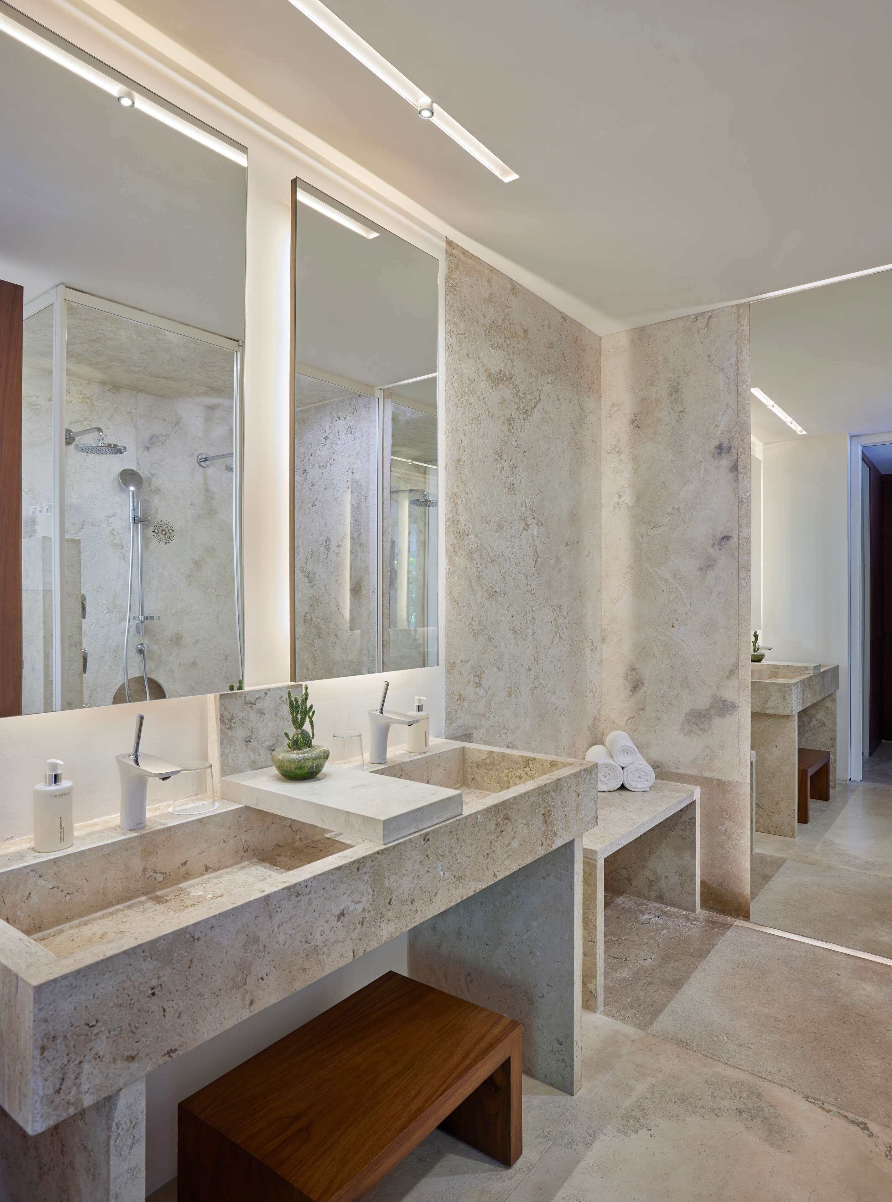 A modern bathroom with a large mirror, two sinks, a shower, and a wooden bench.