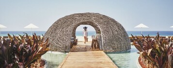 A beachside resort with a thatched archway leading to a pool area, surrounded by lounge chairs and umbrellas.