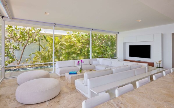 A modern living room with a sectional sofa, a flat-screen TV, and a large window overlooking a pool and greenery.