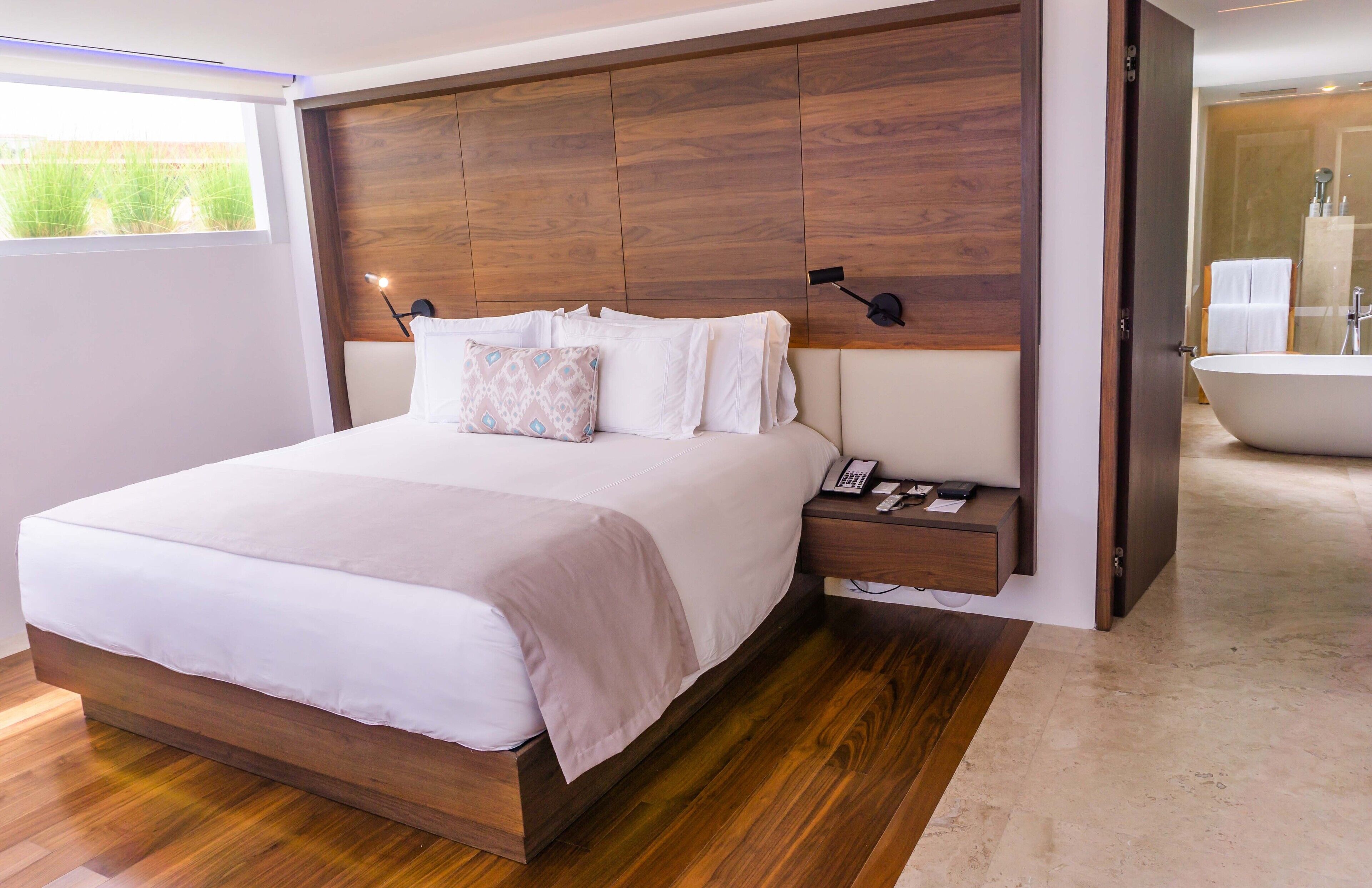 Un dormitorio moderno con cabecera de madera, una cama grande, una mesita de noche con un teléfono, y un baño visible a través de una puerta abierta.