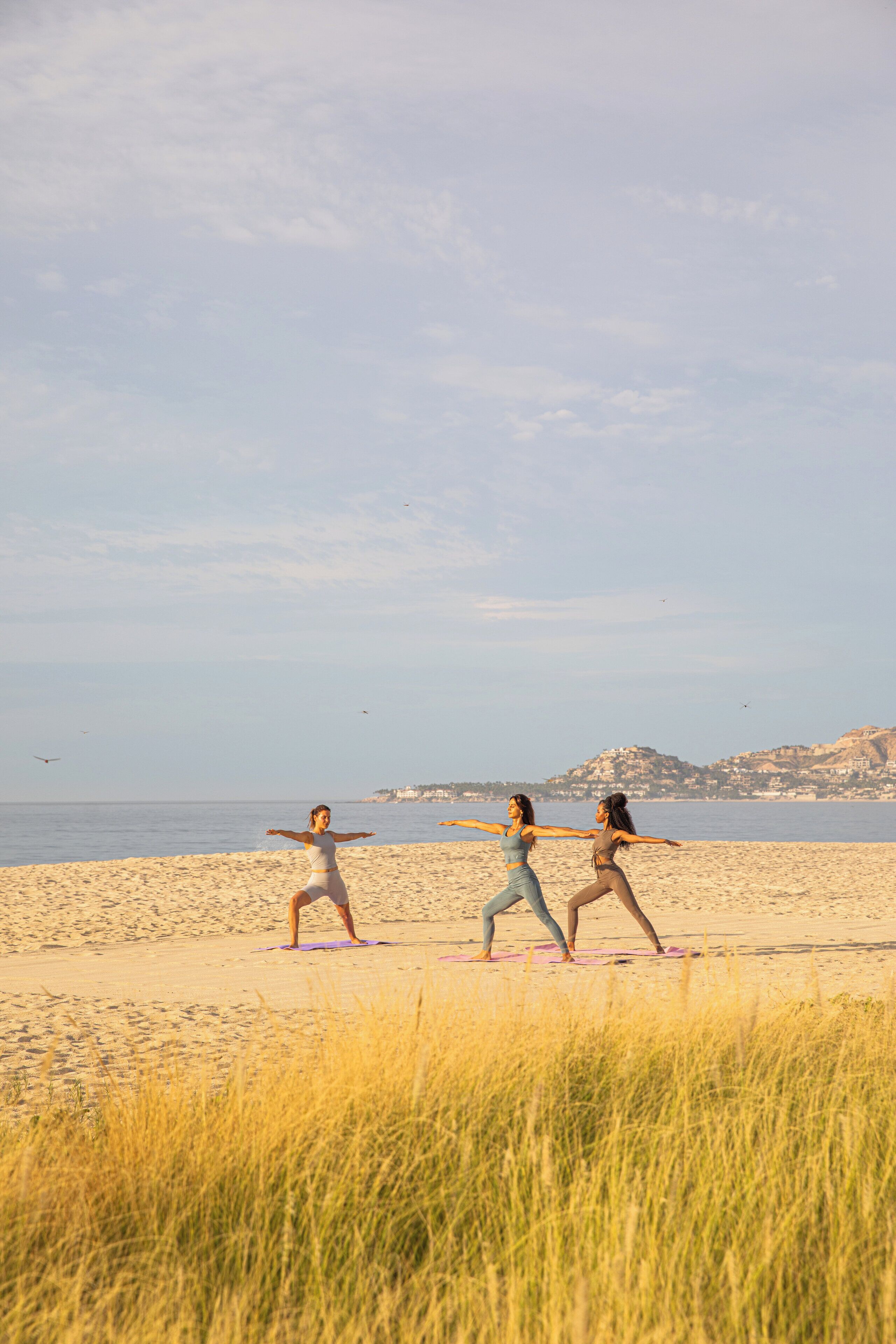 Drie personen beoefenen yoga op een strand, met een heldere lucht en heuvels in de verte.