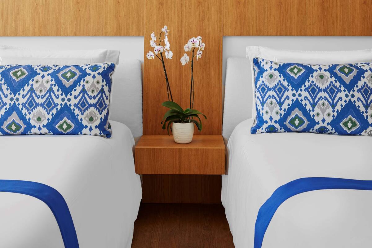 Habitación de hotel con dos camas, cada una con ropa de cama blanca y almohadas estampadas en azul y verde. Entre las camas hay una mesita de noche de madera con una planta de orquídea en maceta.