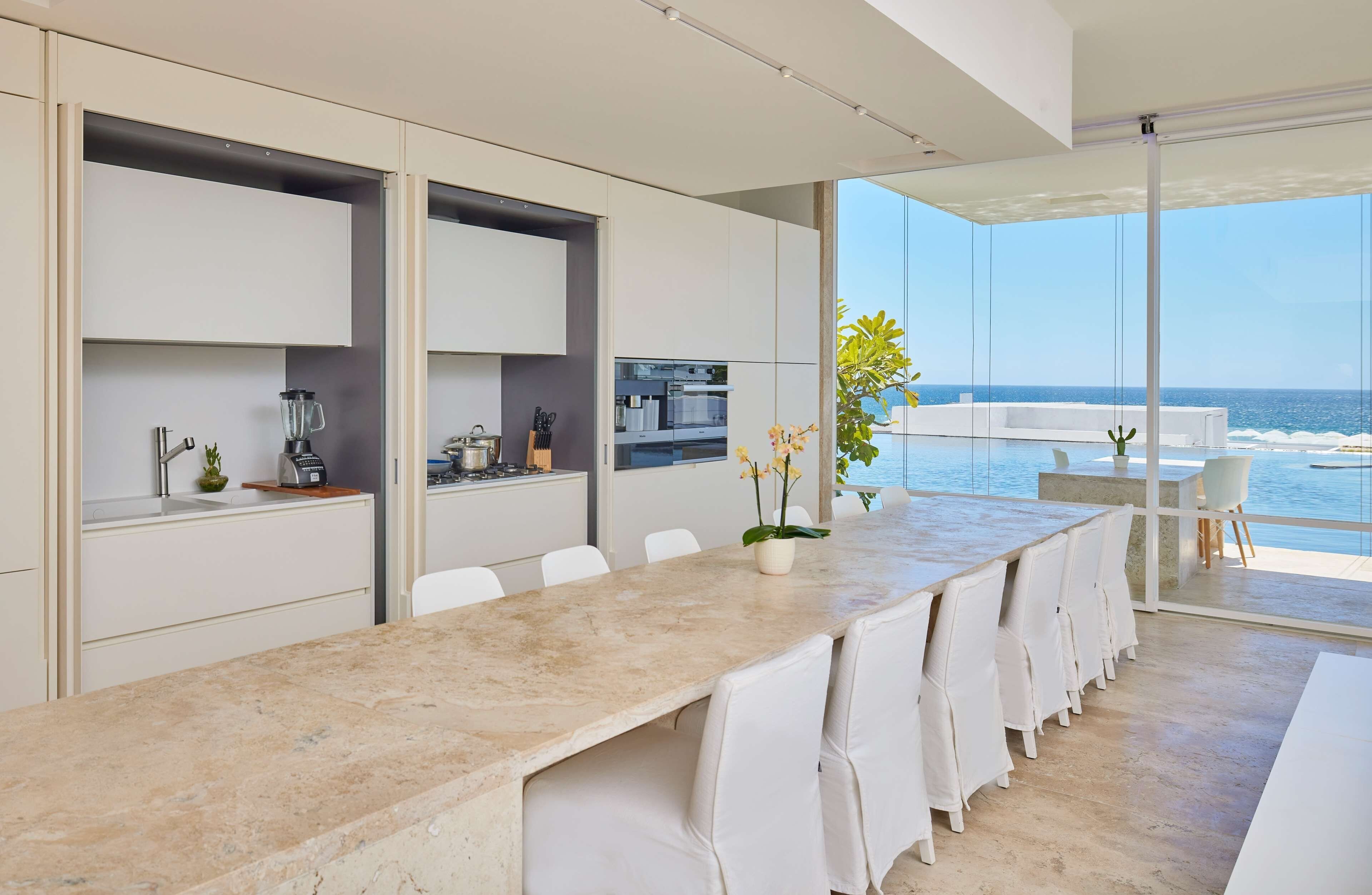 Una cocina moderna con una larga mesa de comedor, sillas blancas y vistas al océano.
