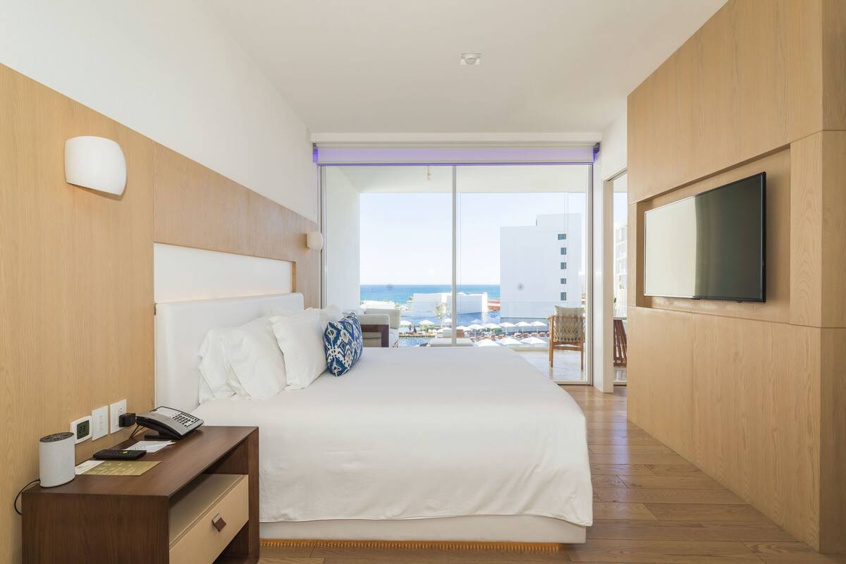 Habitación de hotel con una cama grande, un escritorio con teléfono, un televisor de pantalla plana en la pared y vista al mar a través de amplias ventanas.