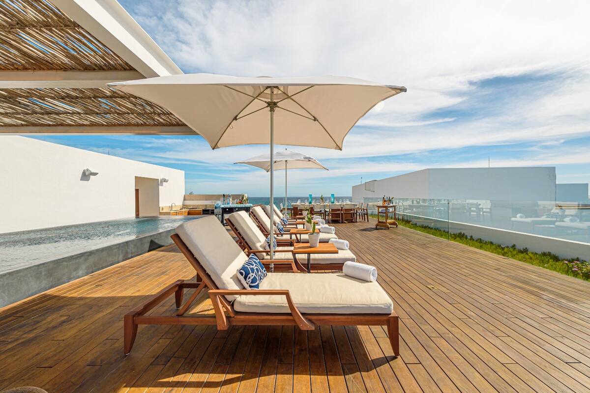 Terraza de madera con sillones de descanso y sombrillas, con vista a una piscina y un paisaje urbano.