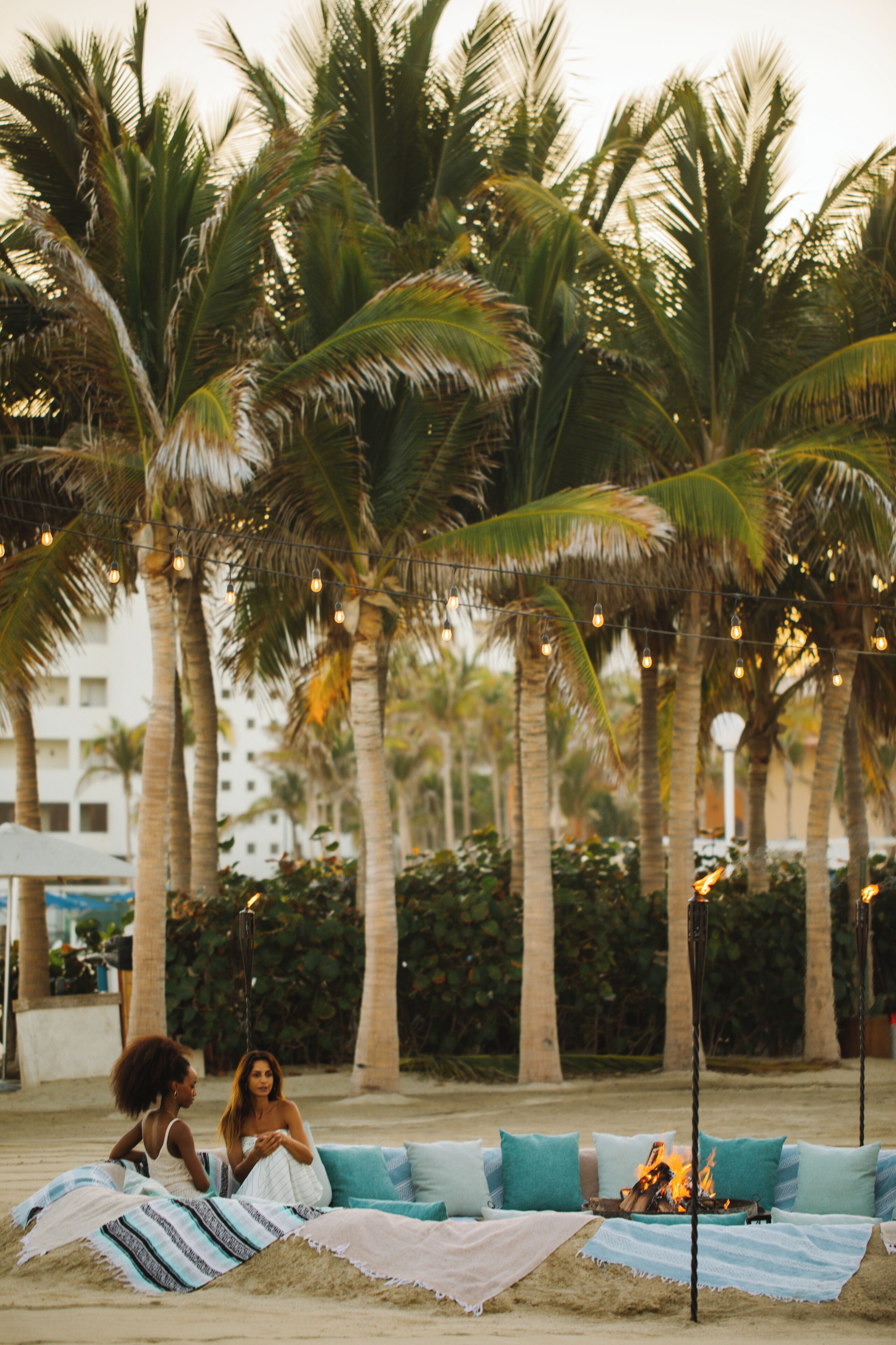 A beach setting with palm trees, a fire pit, and a seating area with cushions.