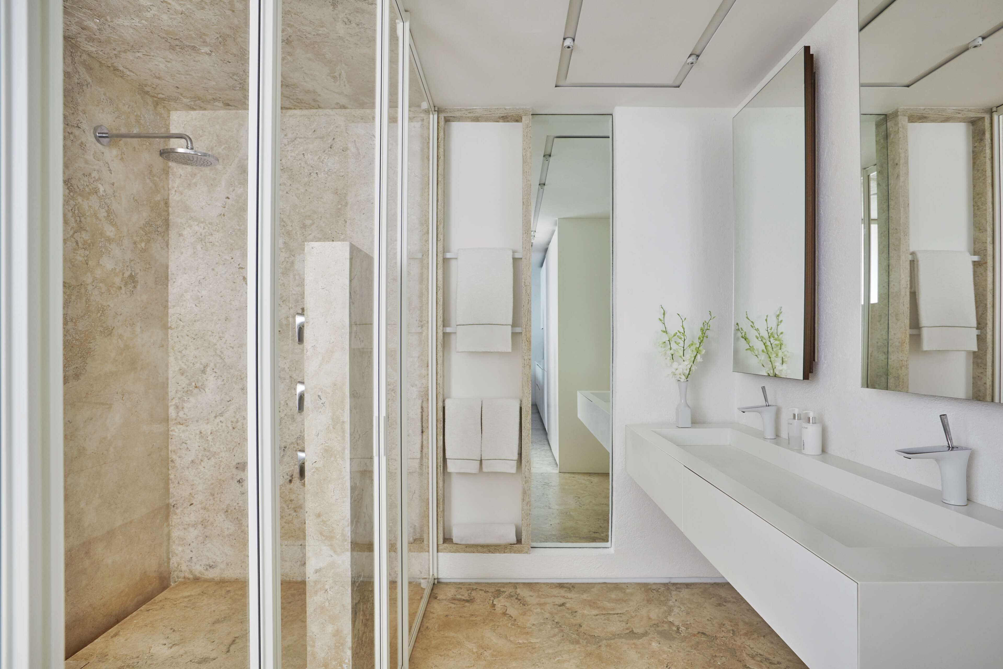 Un baño moderno con cabina de ducha de vidrio, tocador blanco con espejo grande y suelo de mármol.