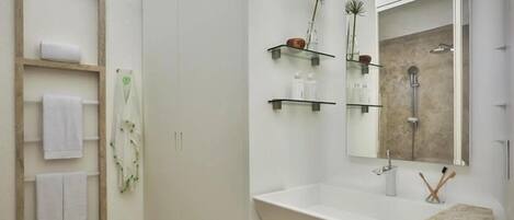 Et moderne badeværelse med et stort spejl, en vask og en bruser. Der er hylder med toiletartikler og håndklæder.