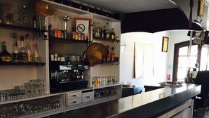 A bar area with shelves of bottles and glasses, a coffee machine, and a counter with stools. - Hôtel Clamador (Saintes-Maries-de-la-Mer)