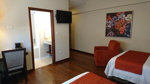 A hotel room with a wooden floor, a bed with a red and white bedspread, a red armchair, a desk with a coffee maker, a TV mounted on the wall, and a painting of flowers. - Hotel Plaza Chihuahua (Chihuahua)