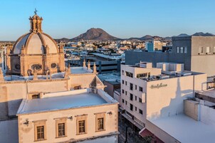 Un paisaje urbano con una cúpula de iglesia destacada, un edificio moderno que dice 'Hotel Plaza' y una montaña al fondo.