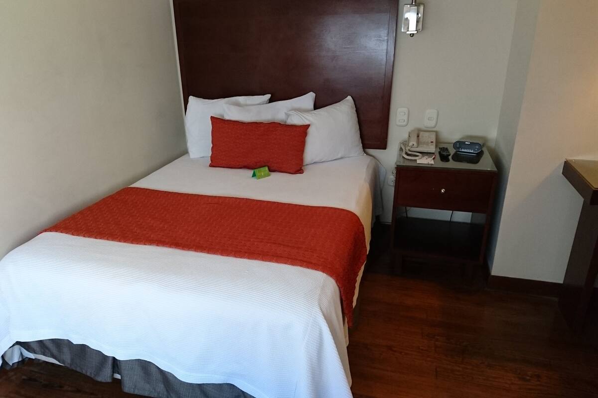 Ein ordentlich bezogenes Bett mit einer roten und weißen Tagesdecke, ein hölzernes Kopfteil, ein Nachttisch mit Telefon und Lampe sowie ein kleiner Holznachttisch.