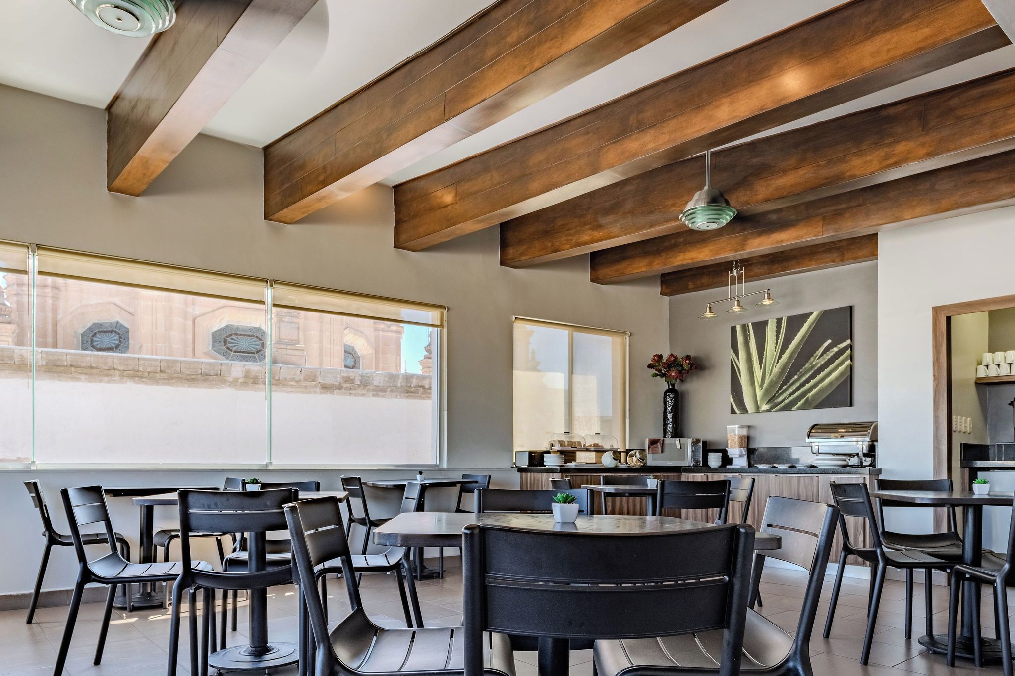 Un comedor moderno con techos altos y vigas a la vista, ventanas amplias con vistas a arquitectura histórica, y una zona de cocina con electrodomésticos de acero inoxidable.