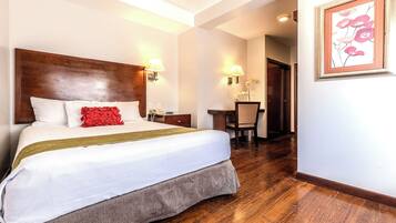 A hotel room with a large bed, a wooden headboard, a desk with a chair, and a framed artwork on the wall.