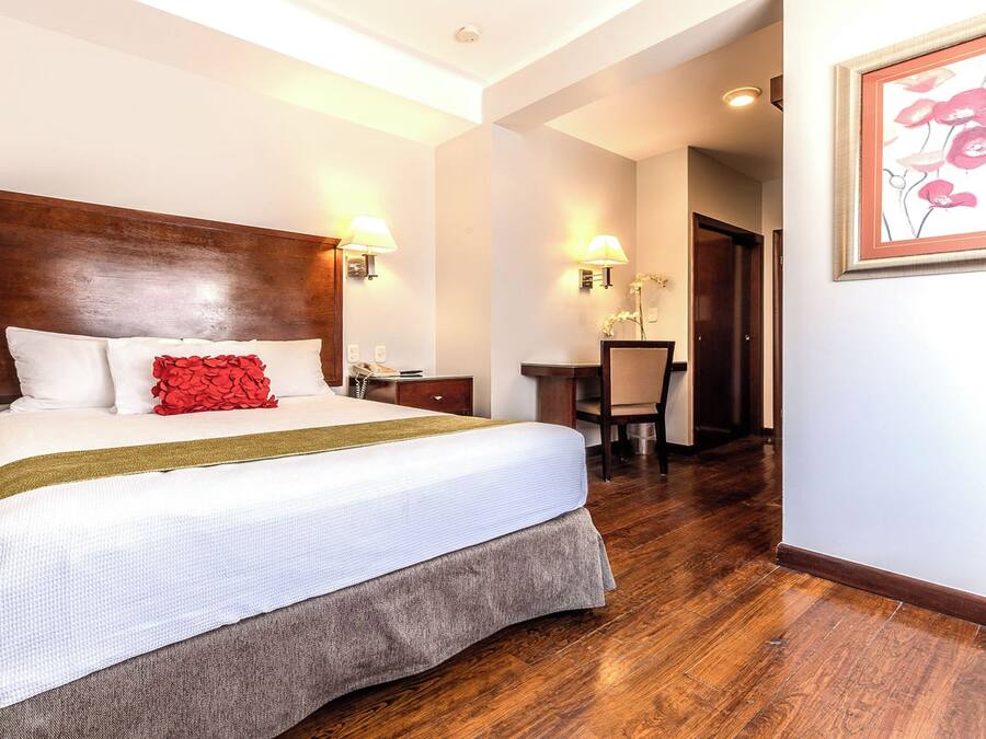 A hotel room with a large bed, a wooden headboard, a desk with a chair, and a framed artwork on the wall.