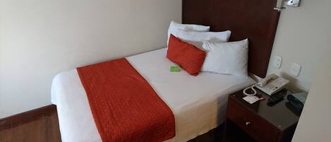 Uma cama bem arrumada com colcha branca e vermelha, cabeceira de madeira e mesa de cabeceira com telefone e controles remotos.