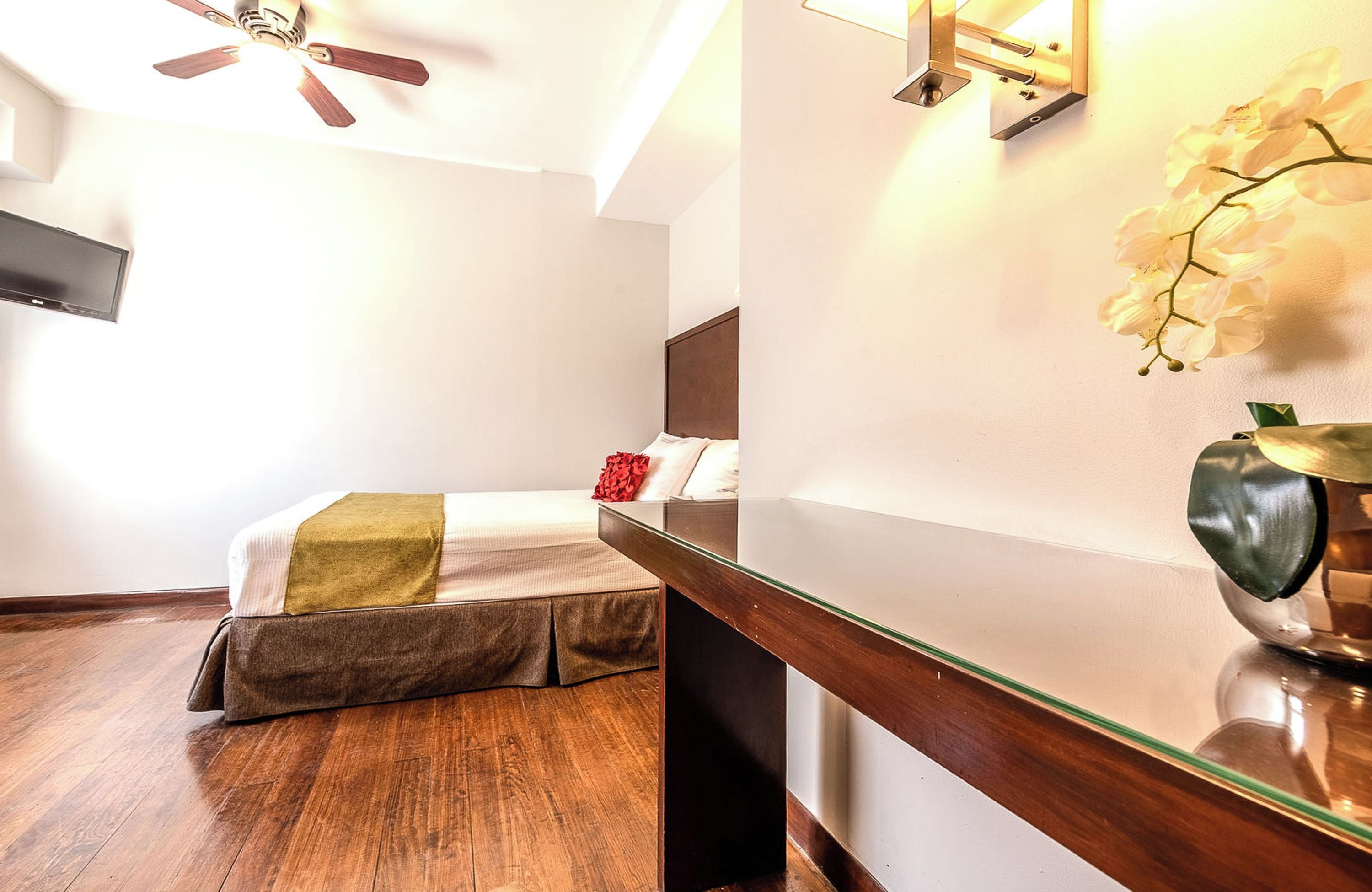 Une chambre d’hôtel avec un lit, un plancher en bois, un ventilateur de plafond, un téléviseur fixé au mur et une orchidée décorative.
